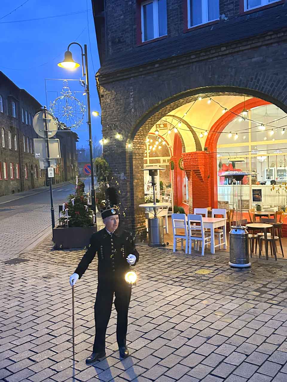 Man dressed as a traditional miner holding a lantern on a cobbled street outside Cafe Byfyj at dusk