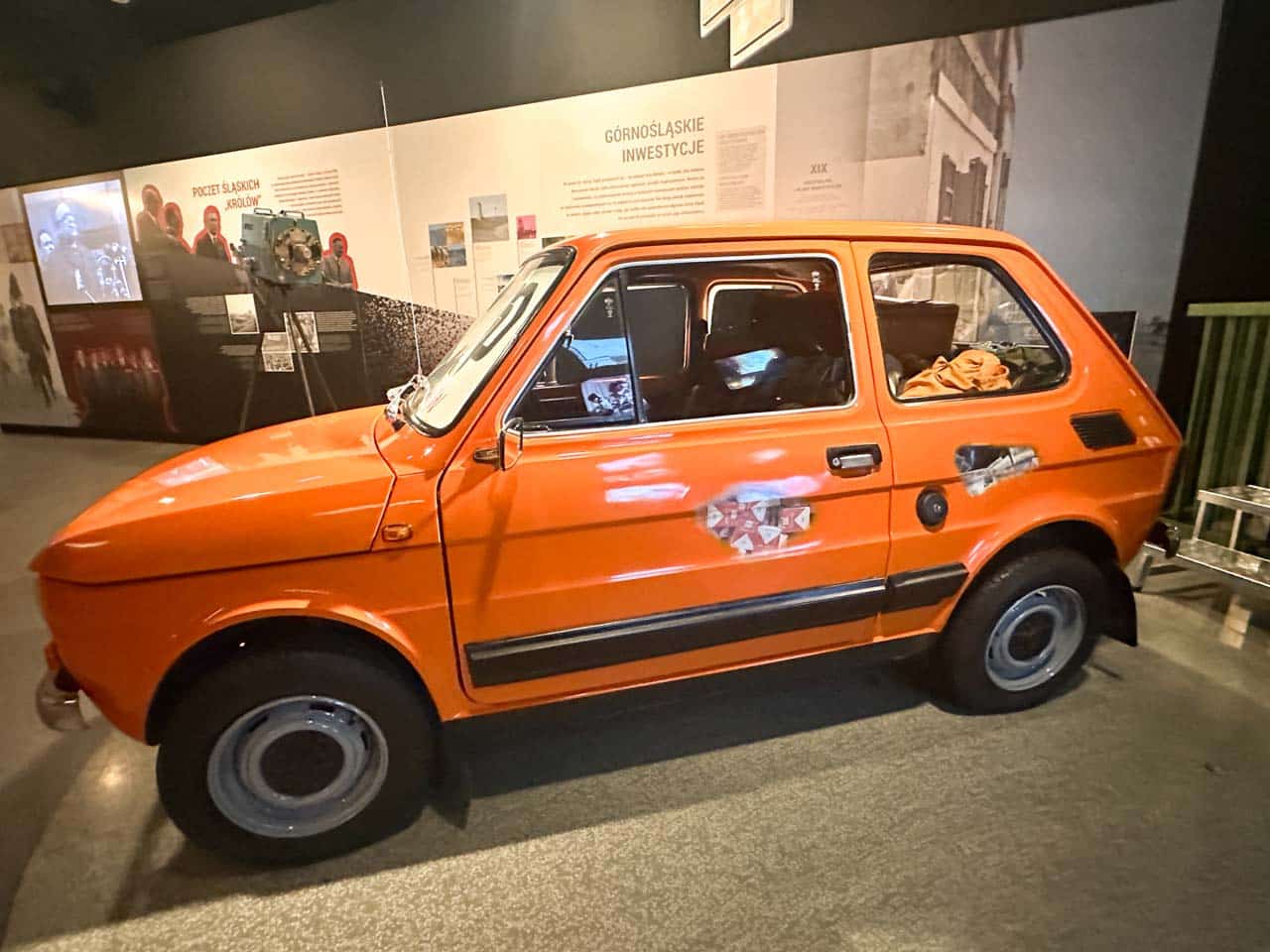 An old orange Fiat 126p car on display inside the Silesian Museum in Katowice, with photos and exhibits in the background