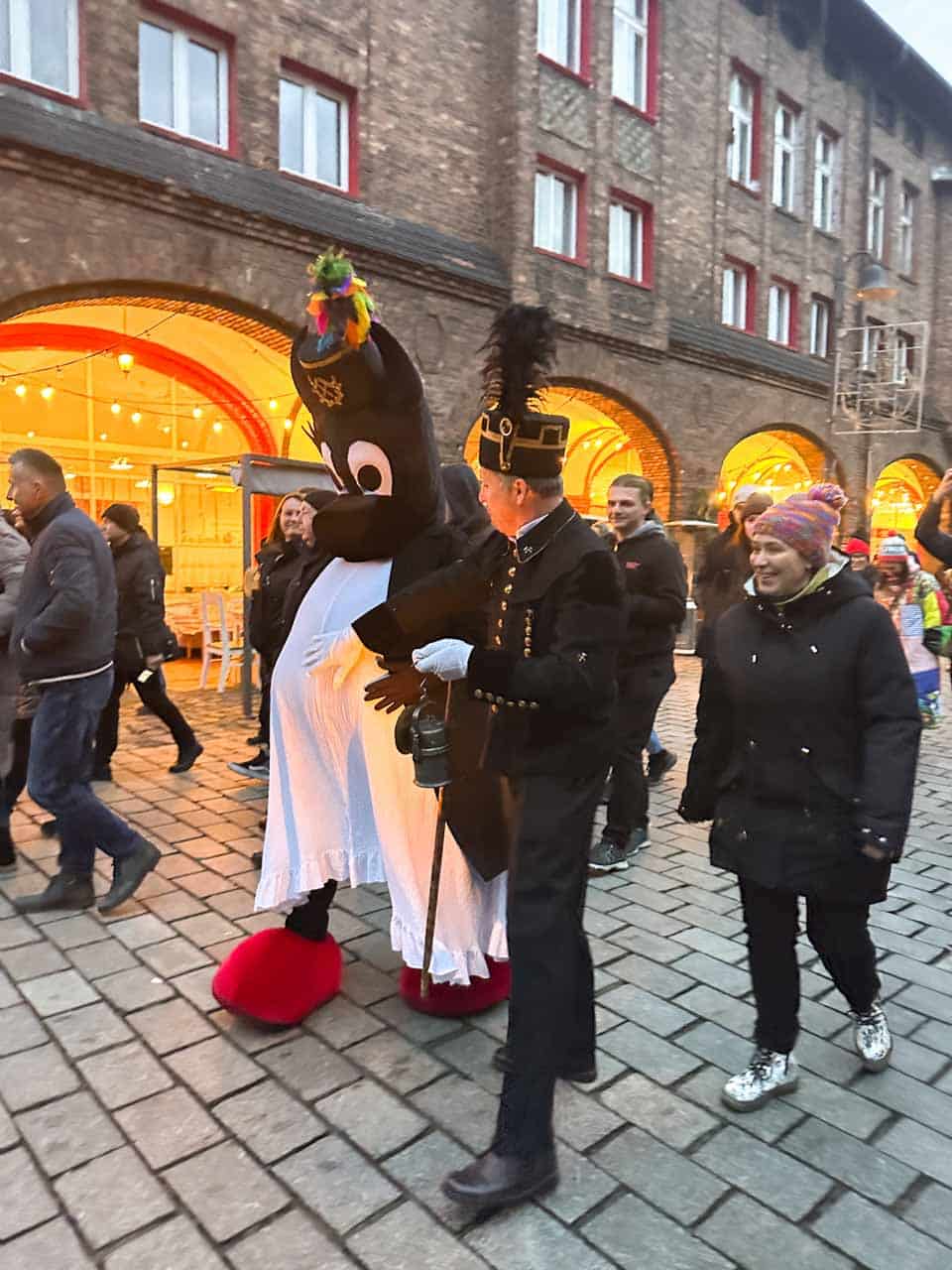 A miner in uniform walking beside a smiling person in a large bebok costume during a Miners' Day event in Nikiszowiec