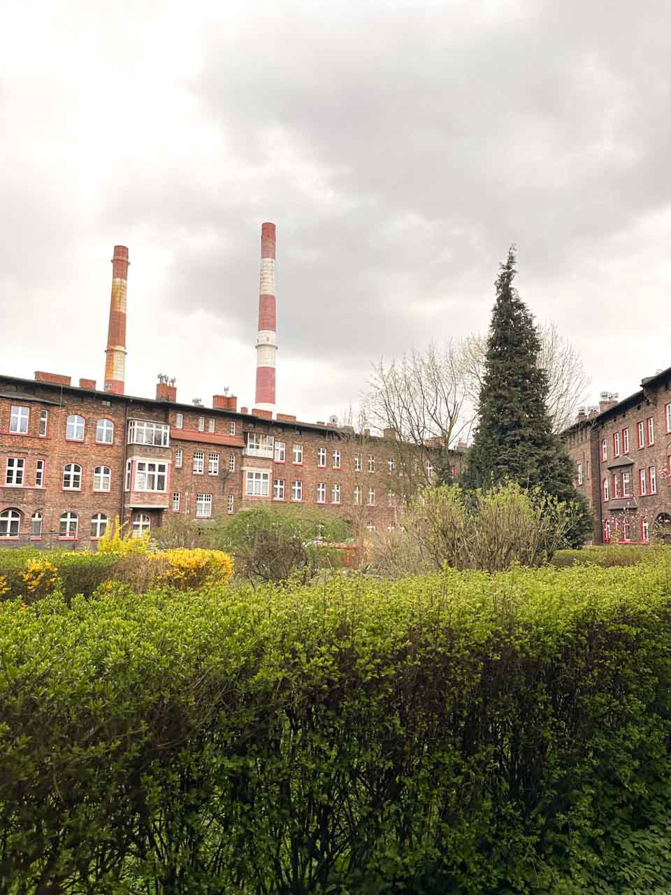 Tall red and white industrial chimneys behind a row of brick buildings and green bushes under a cloudy sky
