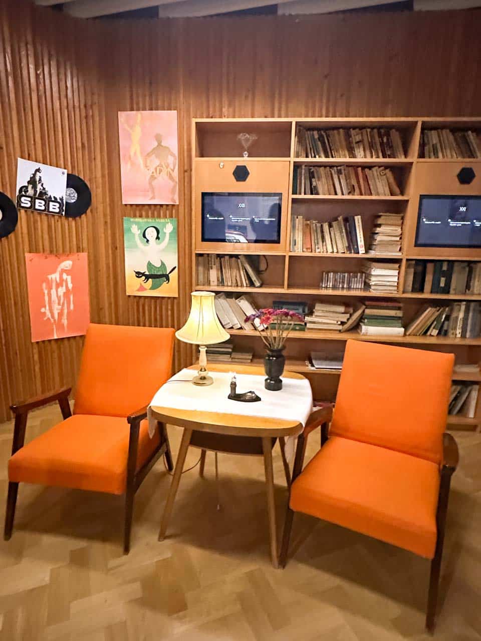 A cosy vintage-style reading nook with two orange armchairs, a small table and lamp, and a wall of books and posters behind