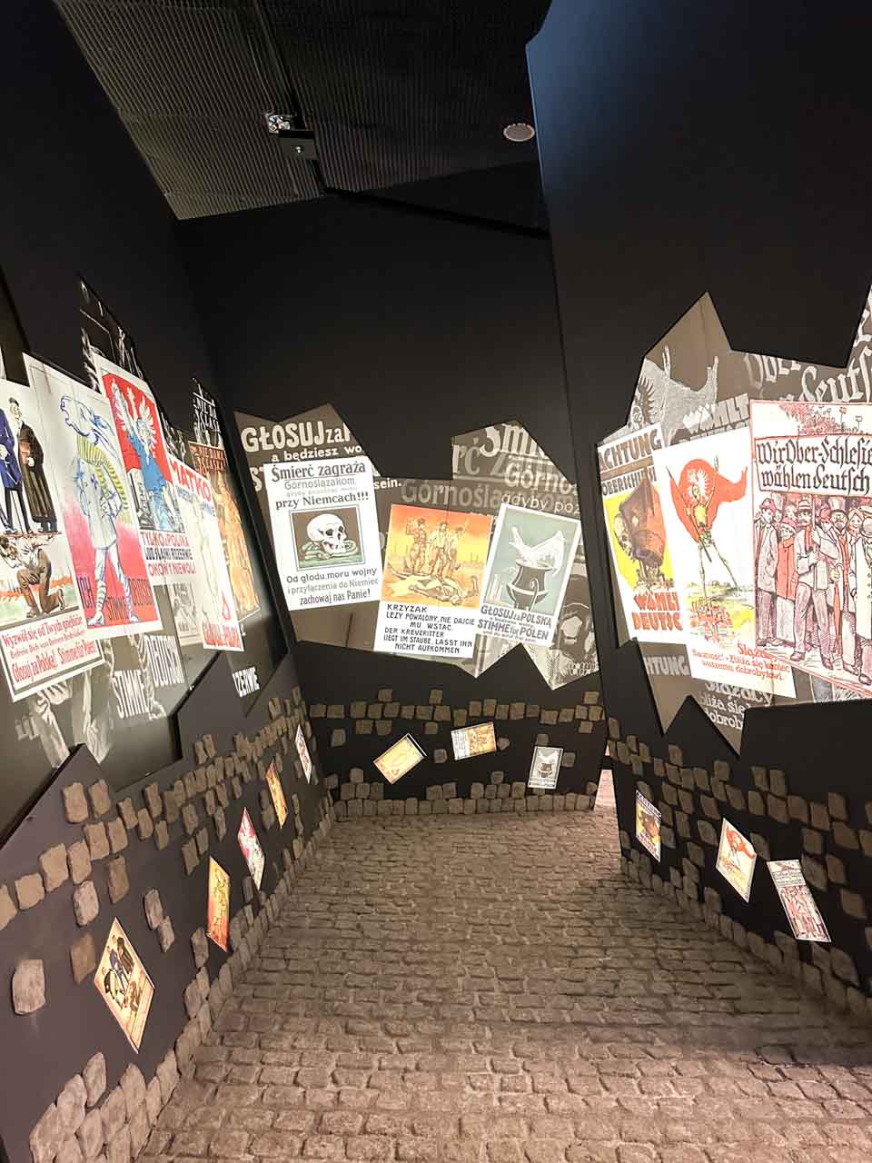 A sloped museum hallway with cobblestone flooring and propaganda posters displayed on black walls