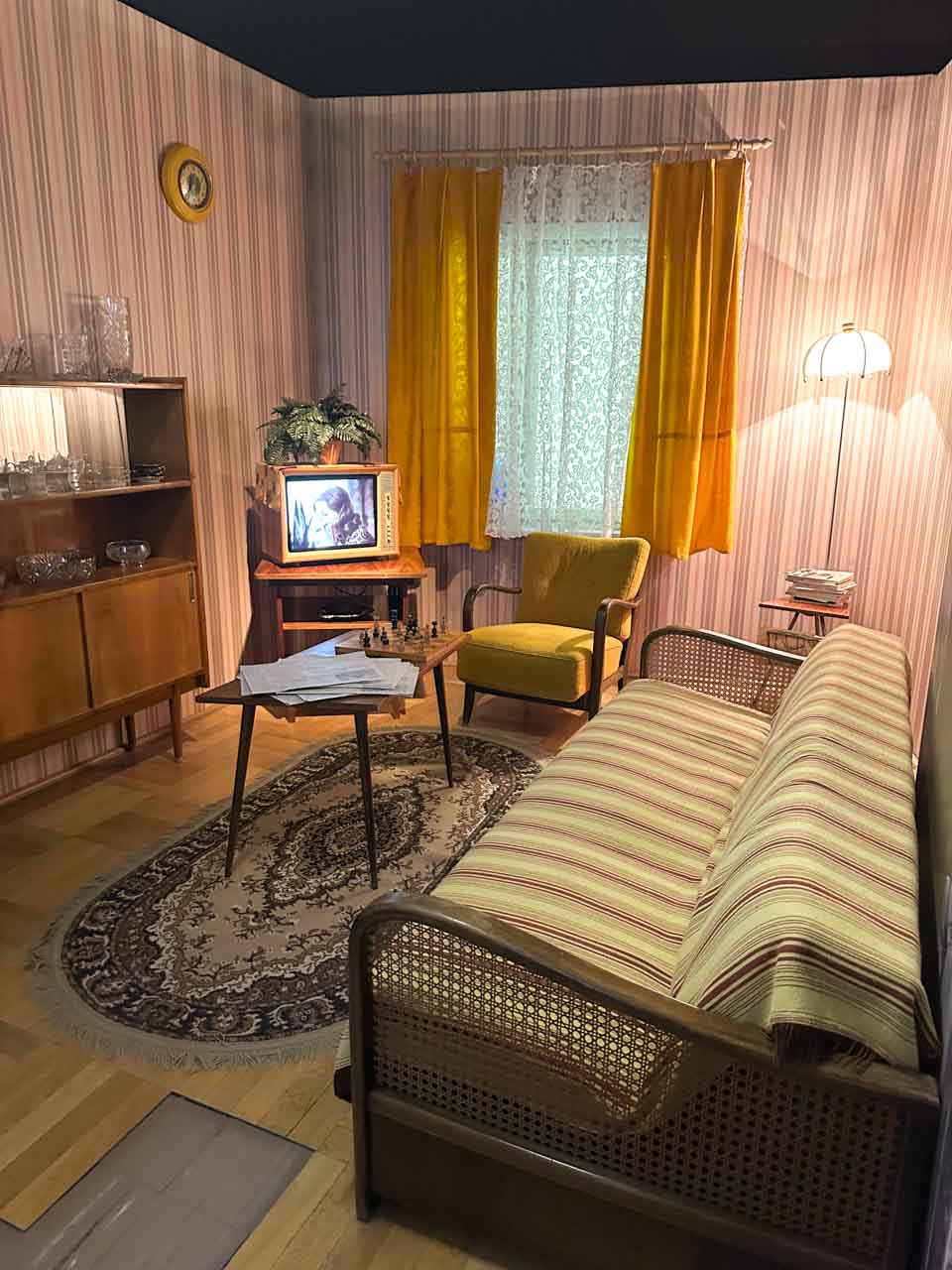 A cosy vintage living room with striped furniture, a lace curtain, and a small TV showing a black and white film at the Silesian Museum in Katowice