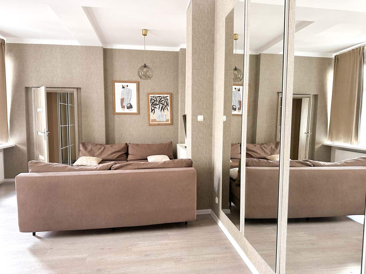 A wide mirror reflecting a light-filled living room with a brown sofa, soft curtains, and two framed artworks on the wall