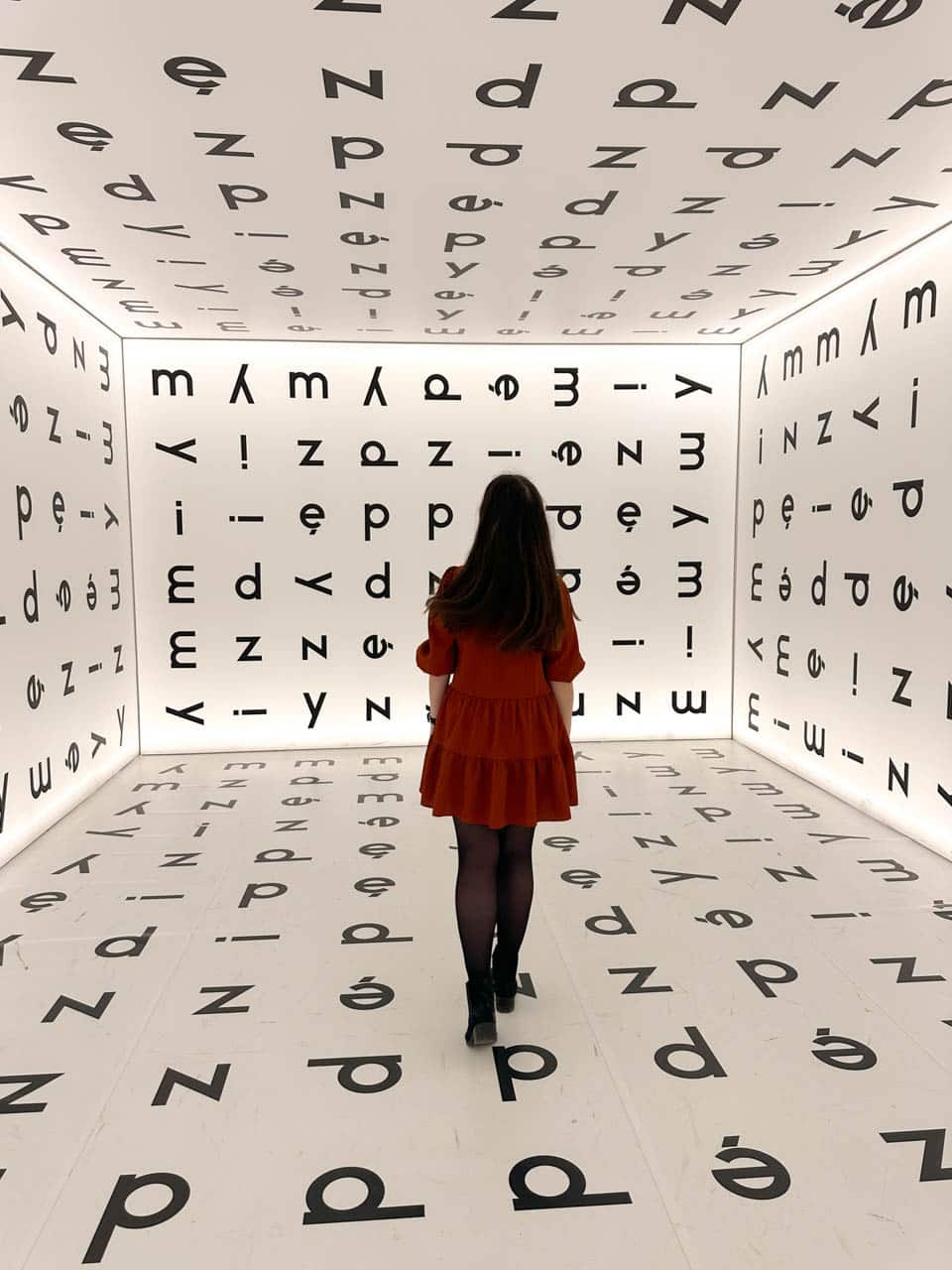 A dark-haired woman in an orange dress walking through a white room covered in oversized black letters on every surface