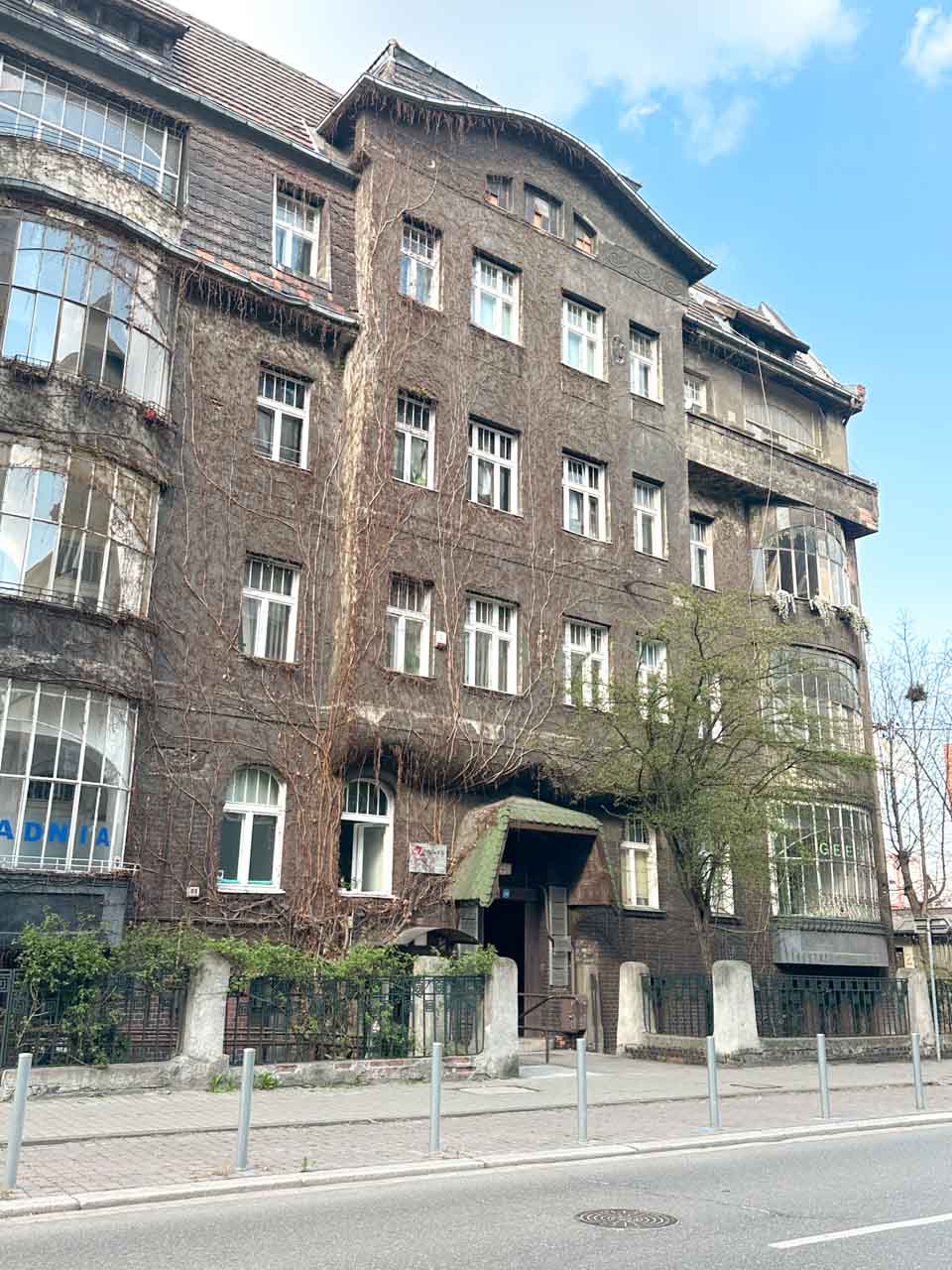A dark ivy-covered building in Katowice with white-framed windows and a green canopy above the entrance