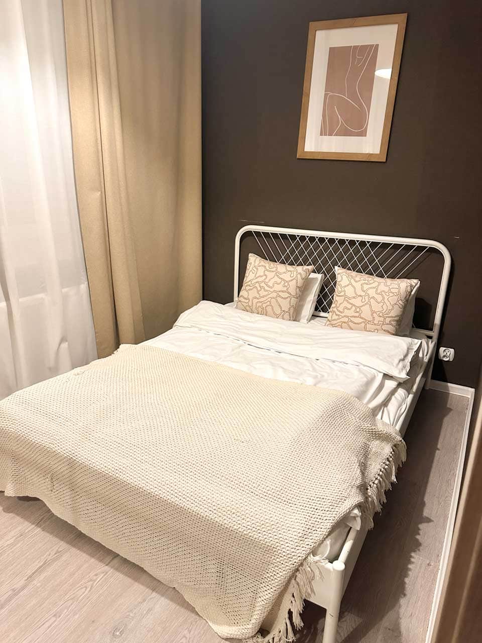 A cosy bedroom with a white metal bed, soft neutral bedding, and abstract art on a dark brown wall