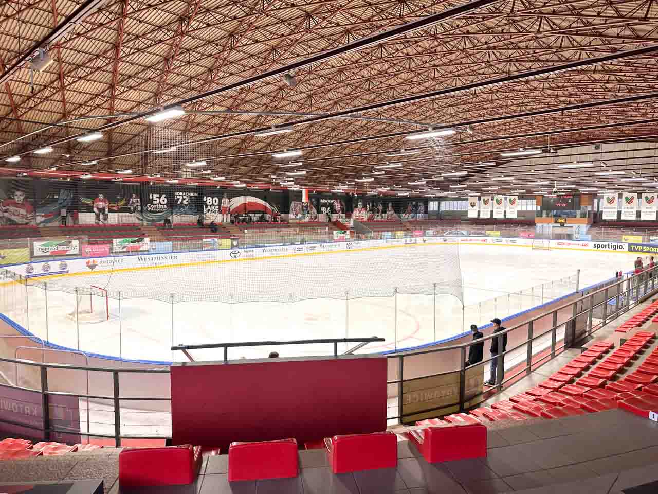 An empty indoor ice hockey arena with red seats, banners on the walls, and a netted rink in the centre