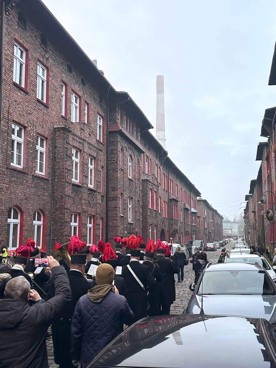 A marching band in black mining uniforms and red plumes parading down a cobbled street lined with red-brick buildings in the Nikiszowiec district of Katowice