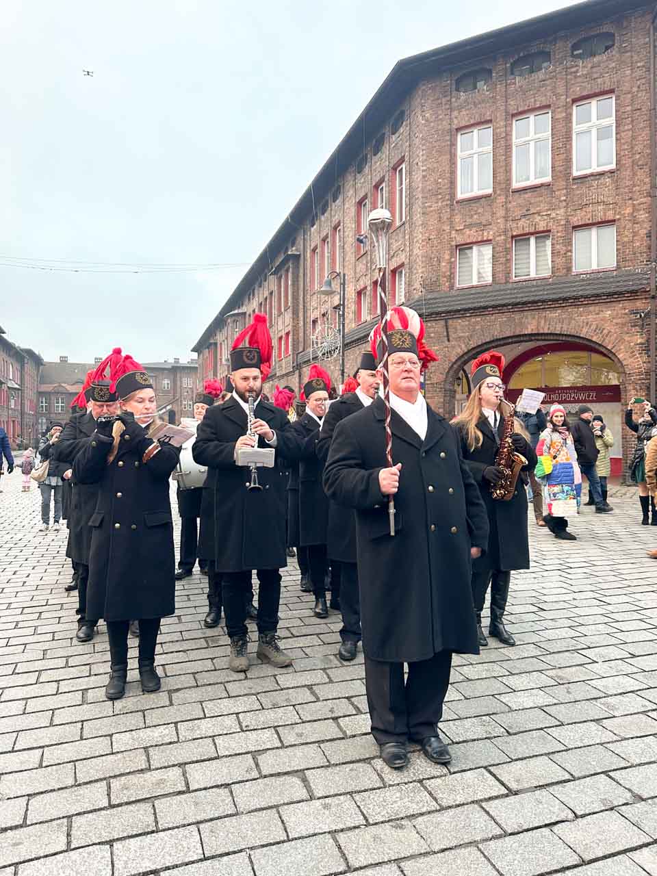 A brass band playing in front of red-brick buildings in the Nikiszowiec district of Katowice, with the leader holding a tall ceremonial baton