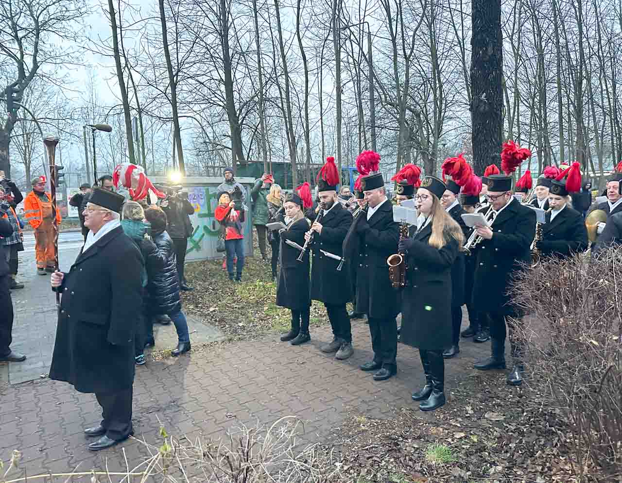 A brass band in black mining uniforms performing outdoors by a path, surrounded by bare trees and people taking photos