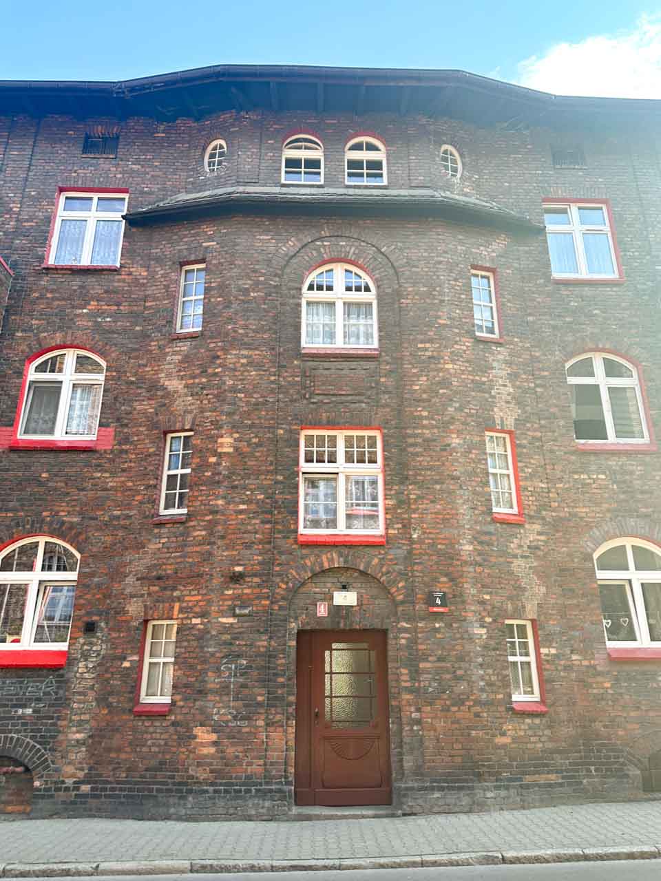 A red-brick tenement building with arched windows and a dark wooden door, framed by a clear blue sky