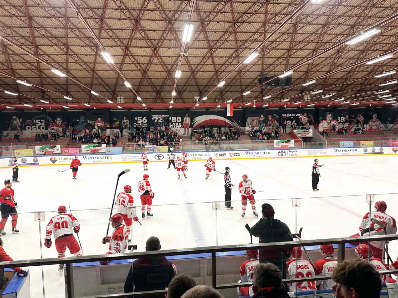 A live ice hockey match with players in red and white jerseys on the rink, and fans watching from the stands