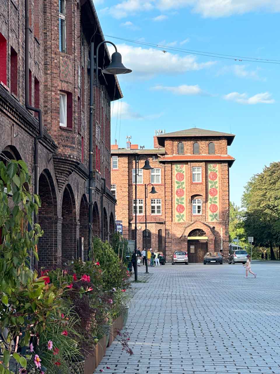 A cobbled square lined with brick buildings, with colourful flower boxes and a wall decorated with red floral designs in the distance
