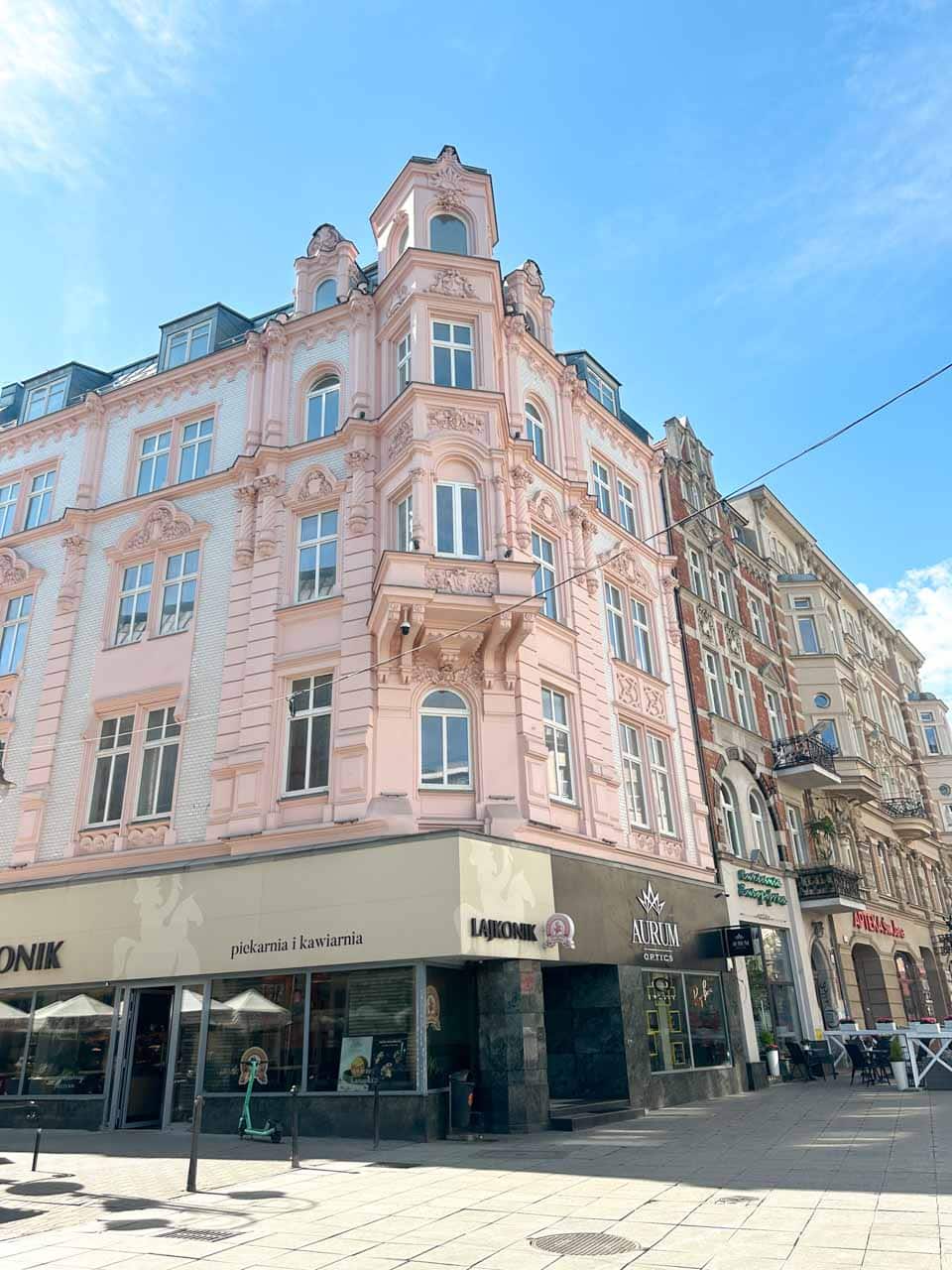 A grand pink building with ornate details and bay windows, housing a bakery and optician at street level