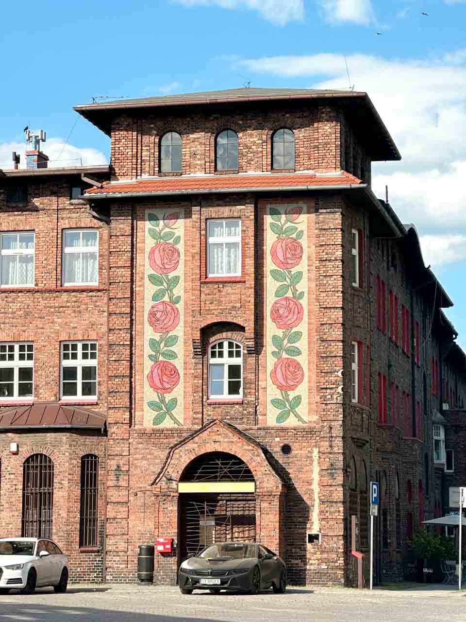 A red brick building with large painted rose patterns on the front, with a black sports car parked outside