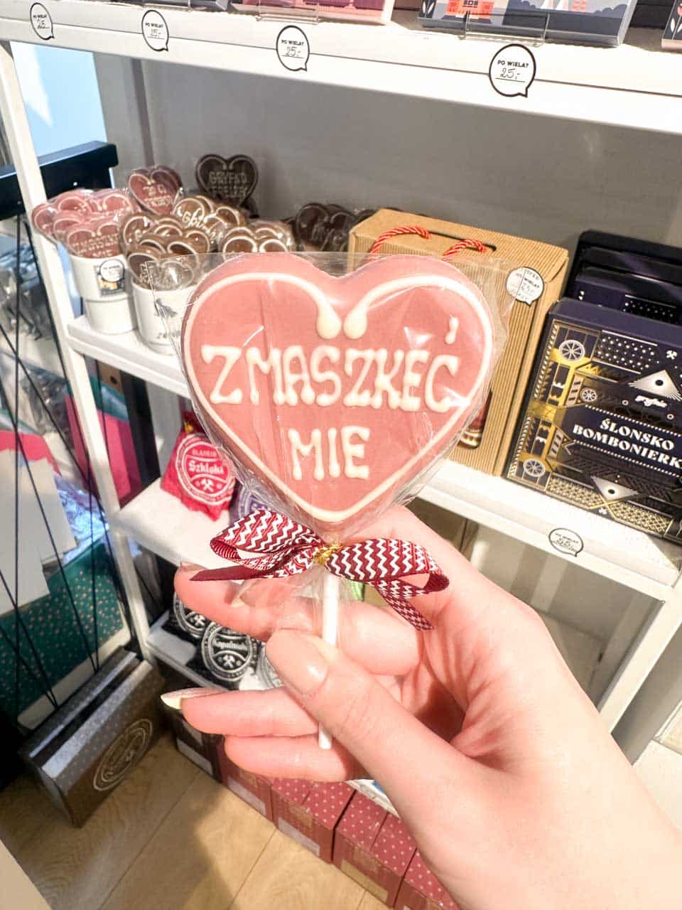 A pink heart-shaped lollipop with Silesian writing, held in front of a shelf full of regional sweets and gifts