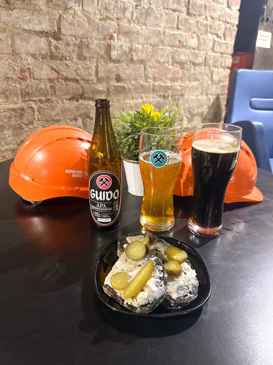 A table with black bread topped with lard and pickles, two beers, and orange mining helmets in the background