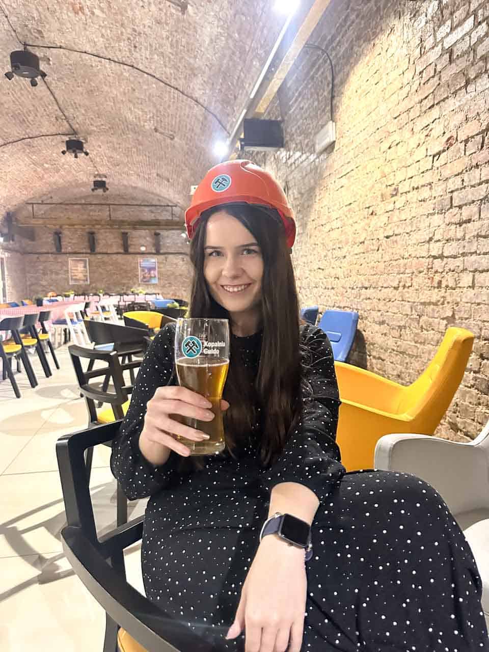 A smiling woman in a polka dot dress wearing a mining helmet and holding a glass of beer at the Guido Coal Mine in Zabrze, Poland