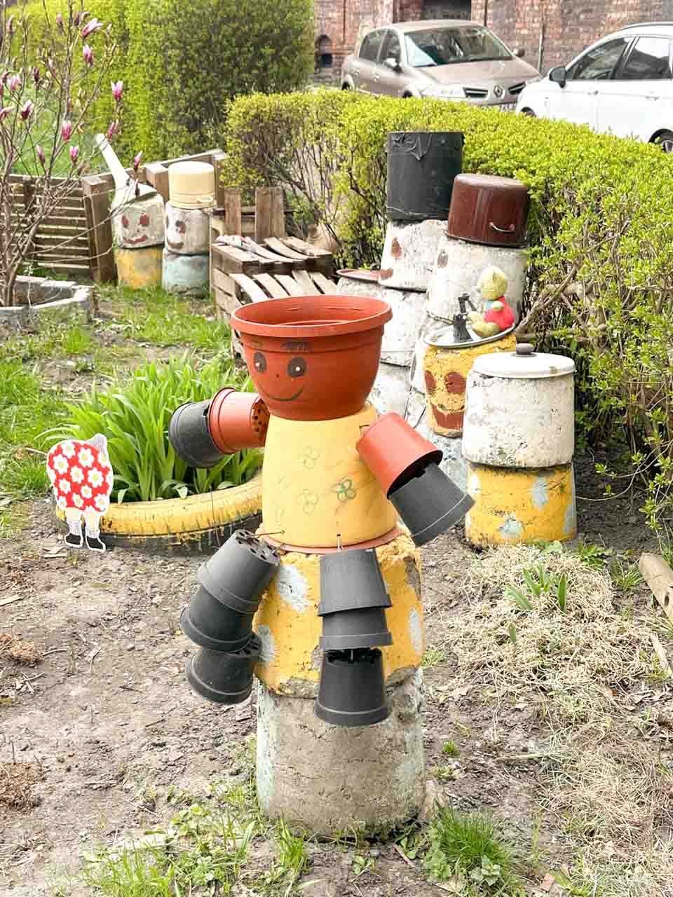 A figure made from flowerpots with a smiley face and arms, standing in a creative DIY garden near painted milk cans in the Nikiszowiec district of Katowice