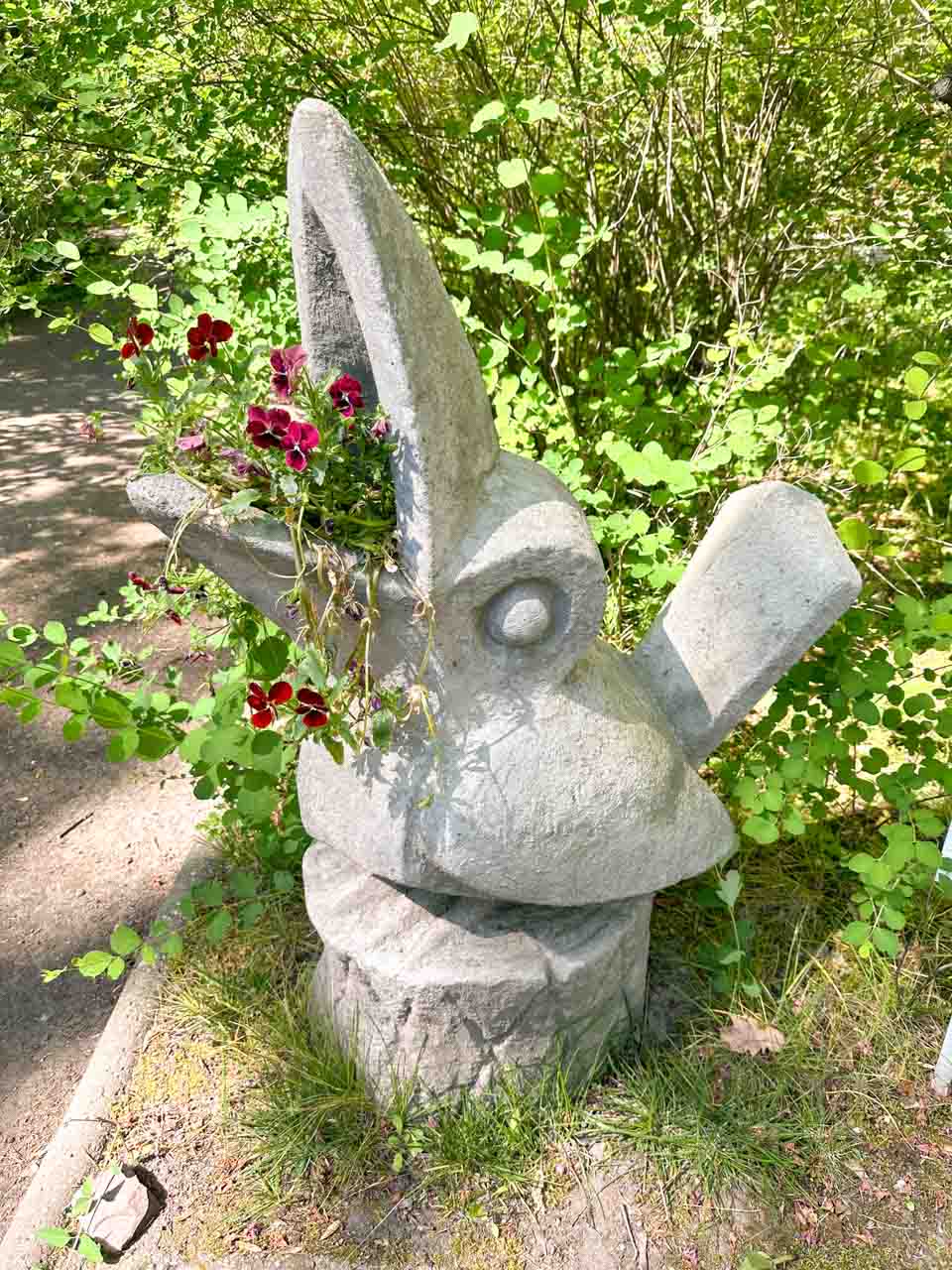 A quirky stone bird sculpture with an open beak, holding dark red flowers like they&rsquo;re being sung out
