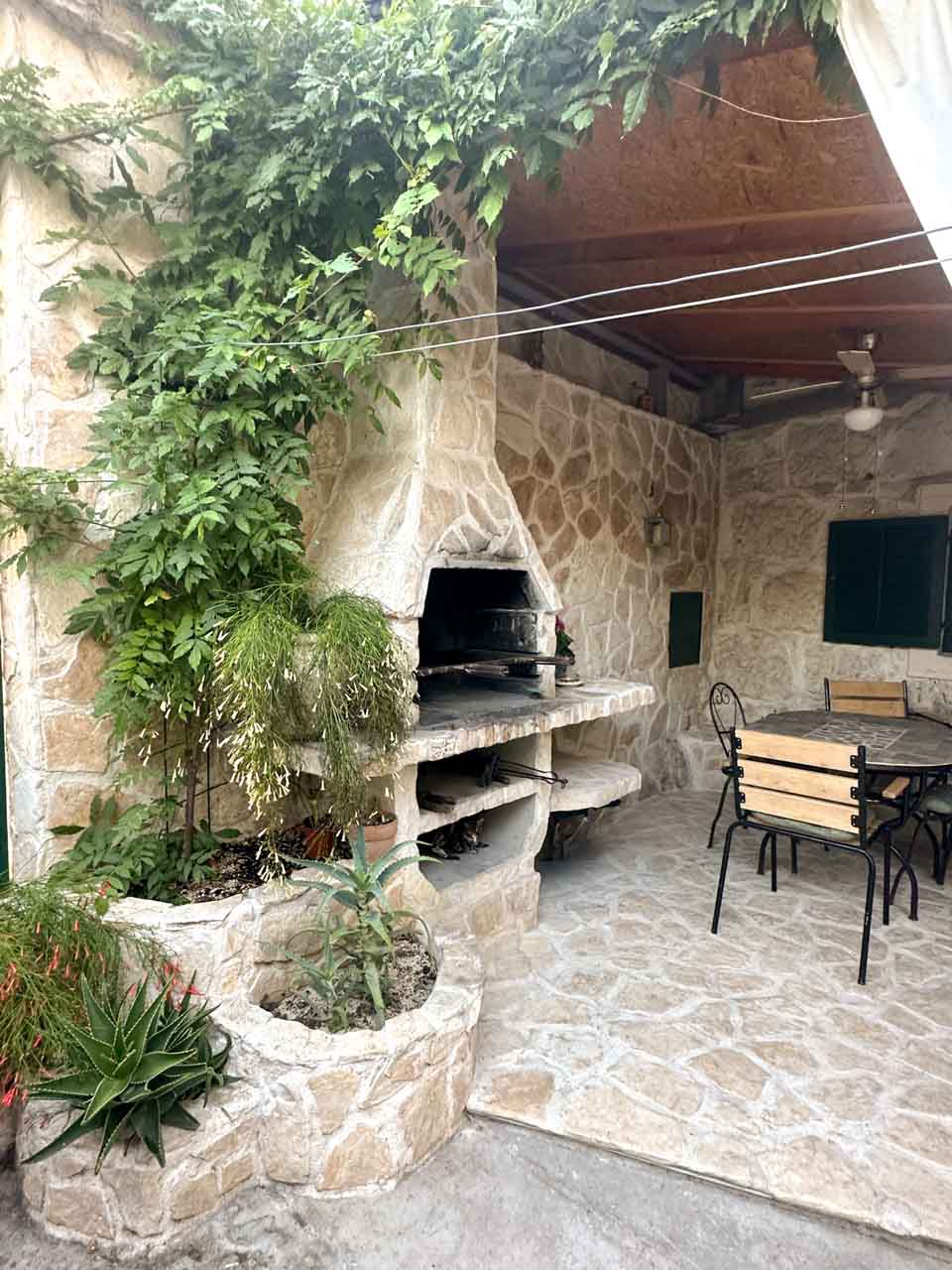 A stone patio with a built-in outdoor grill, leafy green plants, and a table with black metal chairs
