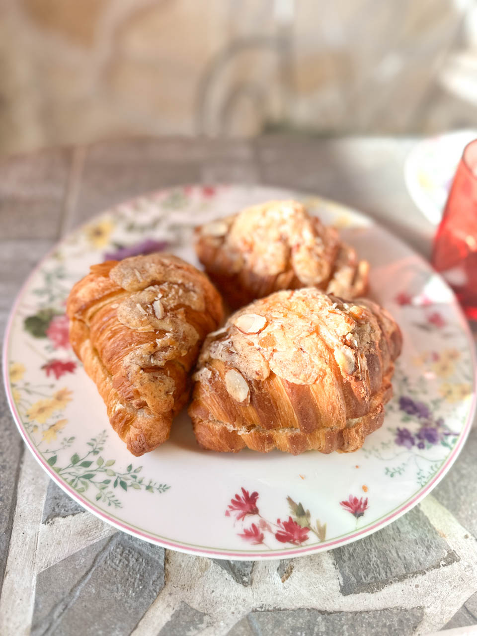Three almond croissants on a pretty floral plate, served at an outdoor stone table