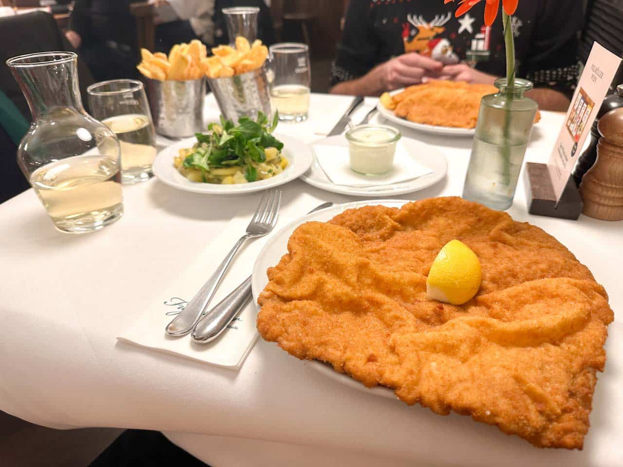 A close-up of a golden schnitzel with a lemon wedge, served with potato salad, chips, and grape juice