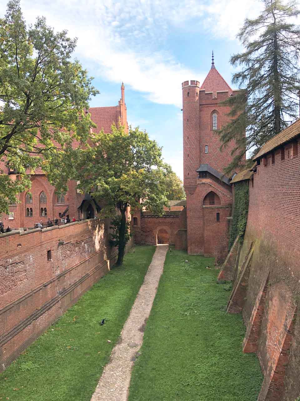Pathway between high red brick walls at Malbork Castle, with a view of the tower and lush greenery