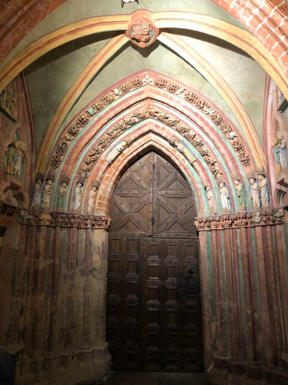 Gothic wooden door with arched carvings, framed by colourful painted figures and floral patterns