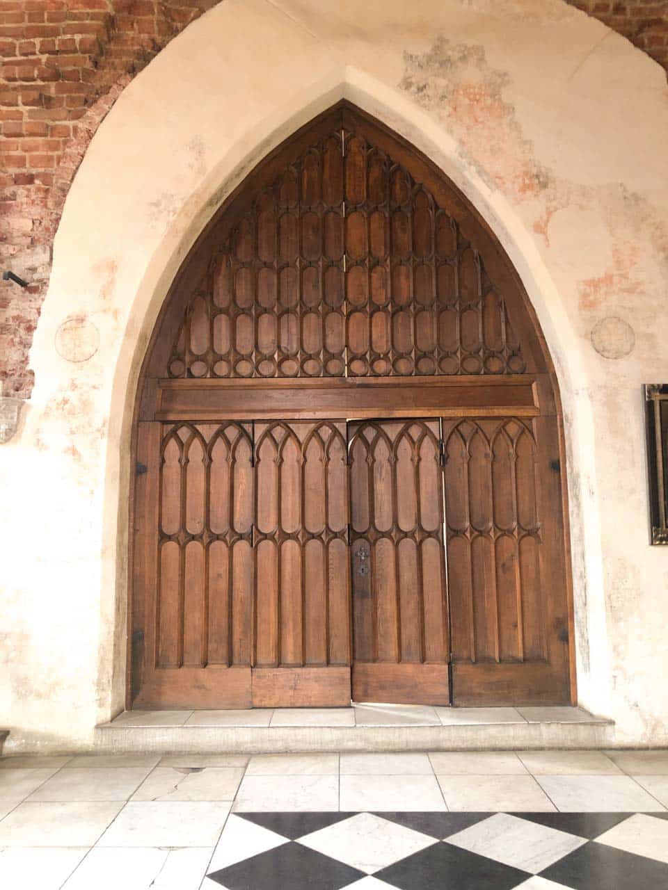 Tall wooden Gothic door with pointed arches, set in a pale stone wall above a black and white tiled floor
