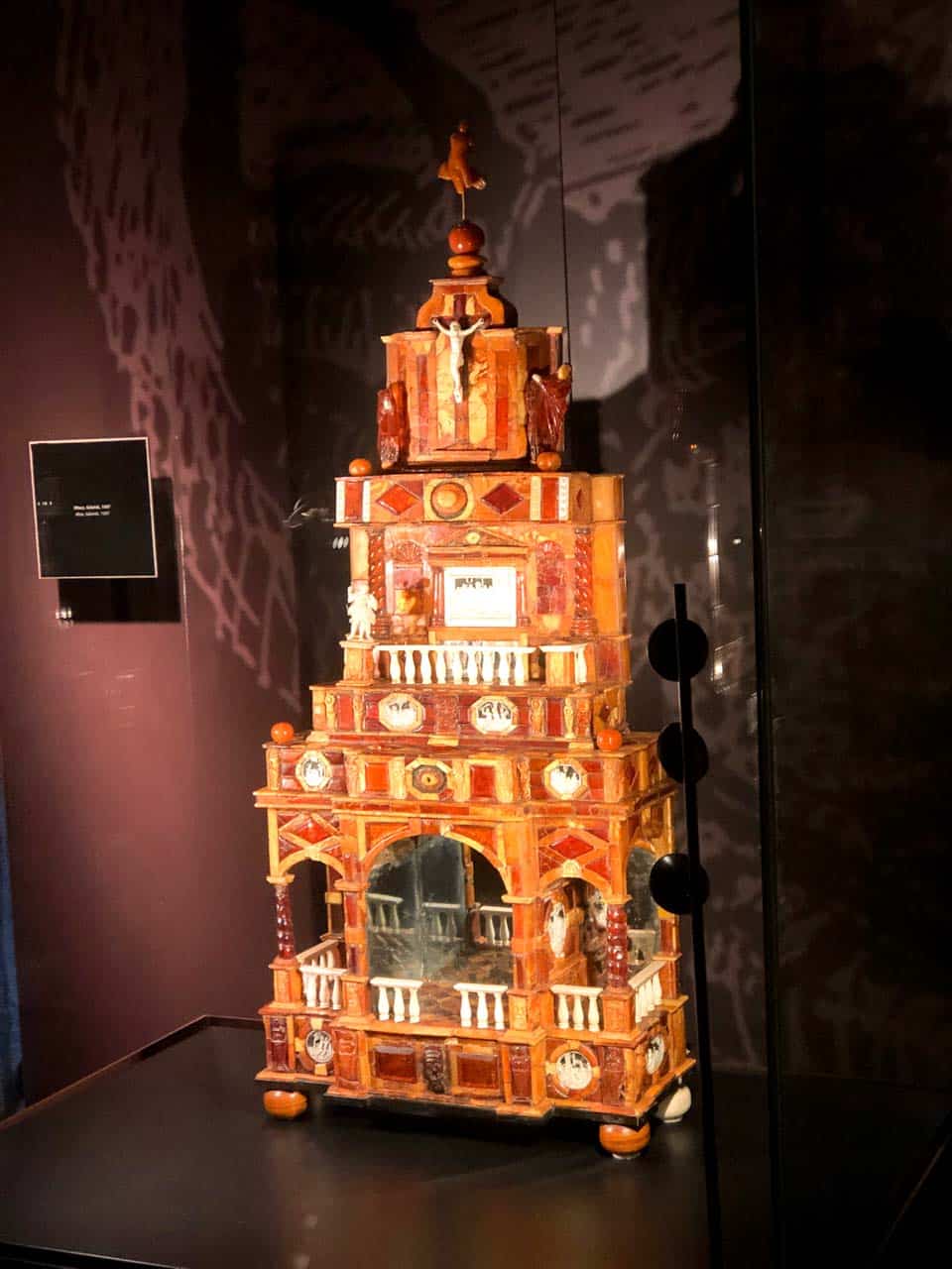 Tall amber cabinet shaped like a miniature chapel, with tiny columns, carvings, and a crucifix at the top, displayed in a glass case