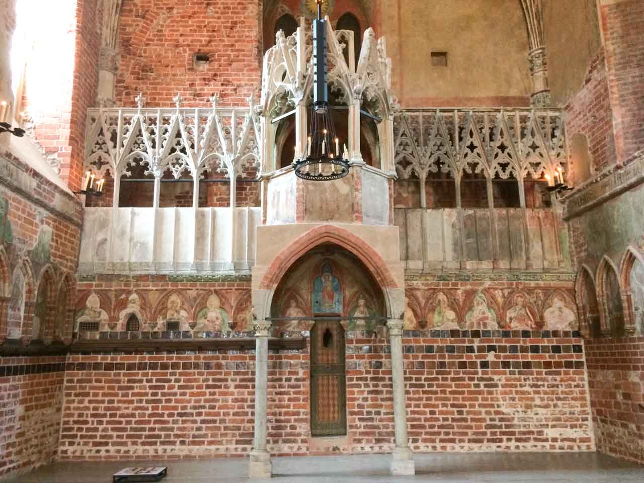 Ornate chapel interior with red brick walls, painted saints, stone carvings, a raised pulpit, and a black iron chandelier