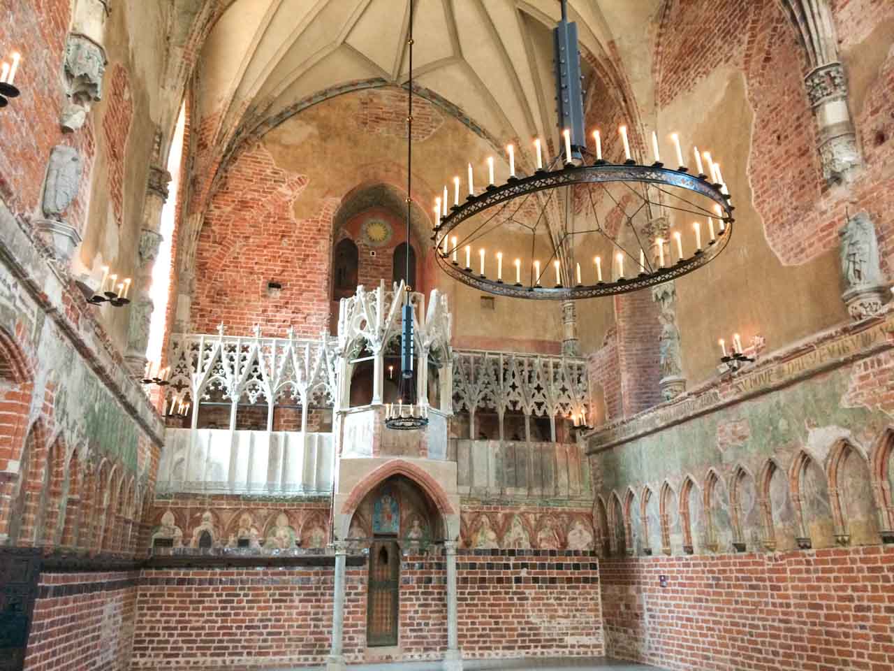 View of a brick-walled chapel inside Malbork Castle with a large round chandelier, tall ceiling, and richly carved pulpit