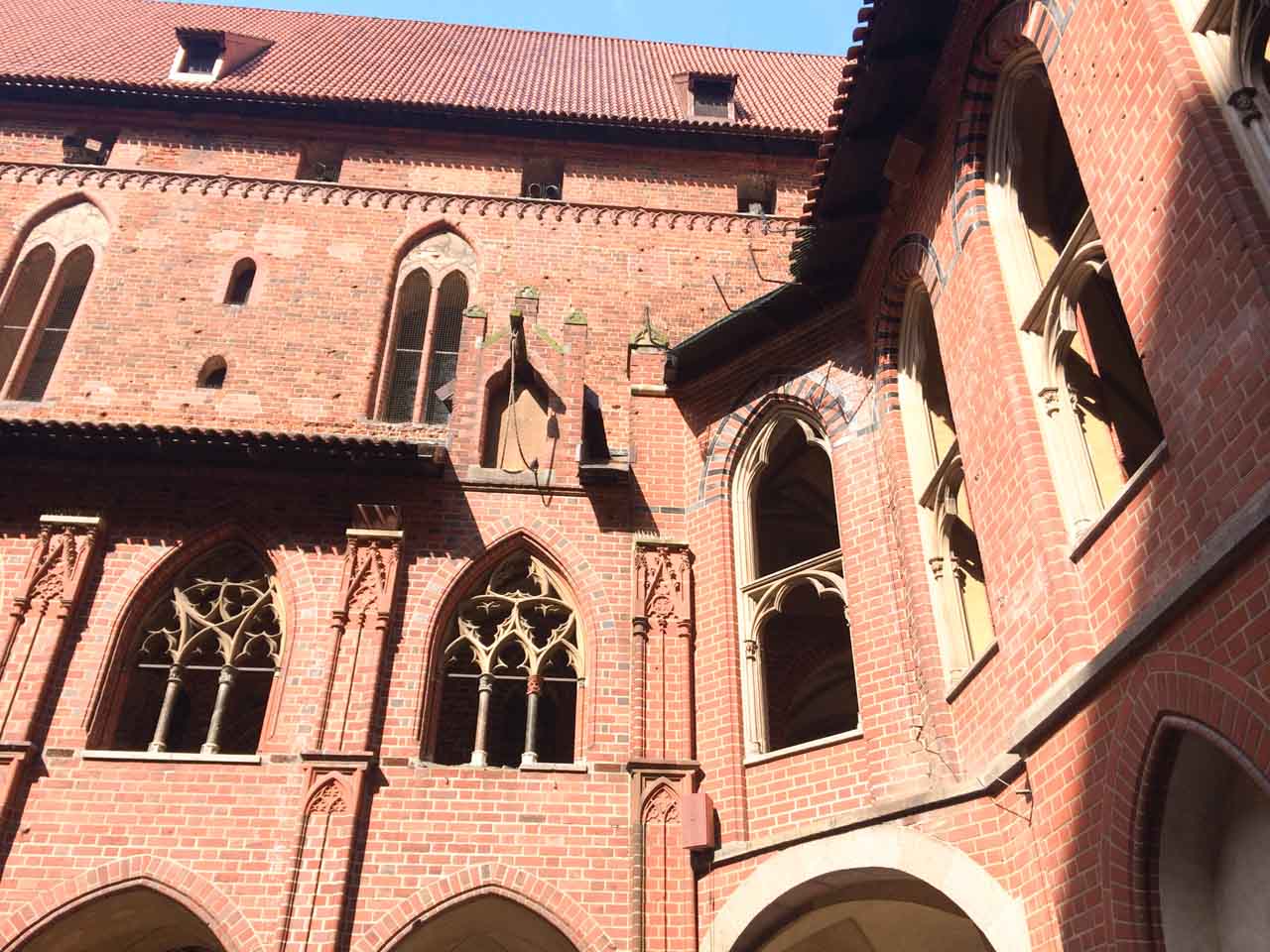 Sunny courtyard of Malbork Castle with red brick walls, gothic windows, and pointed arches around the upper floors