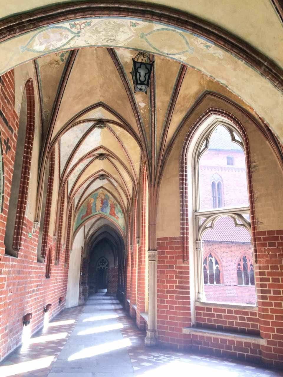 Light-filled cloister of Malbork Castle with vaulted ceilings, tiled floors, and painted details above the arched walkway