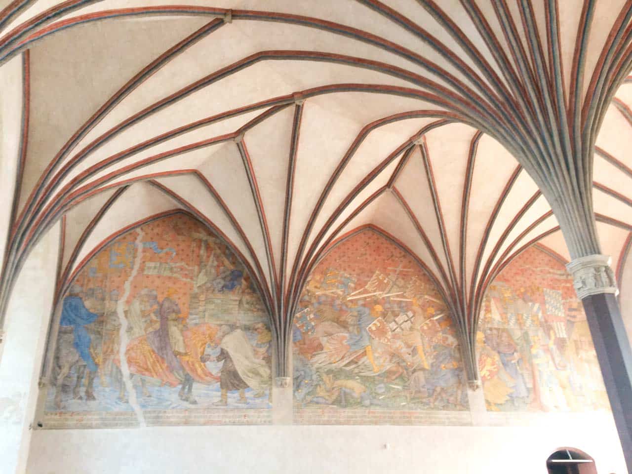 Full fresco wall scene under a ribbed ceiling, showing knights, battle scenes, and royal figures