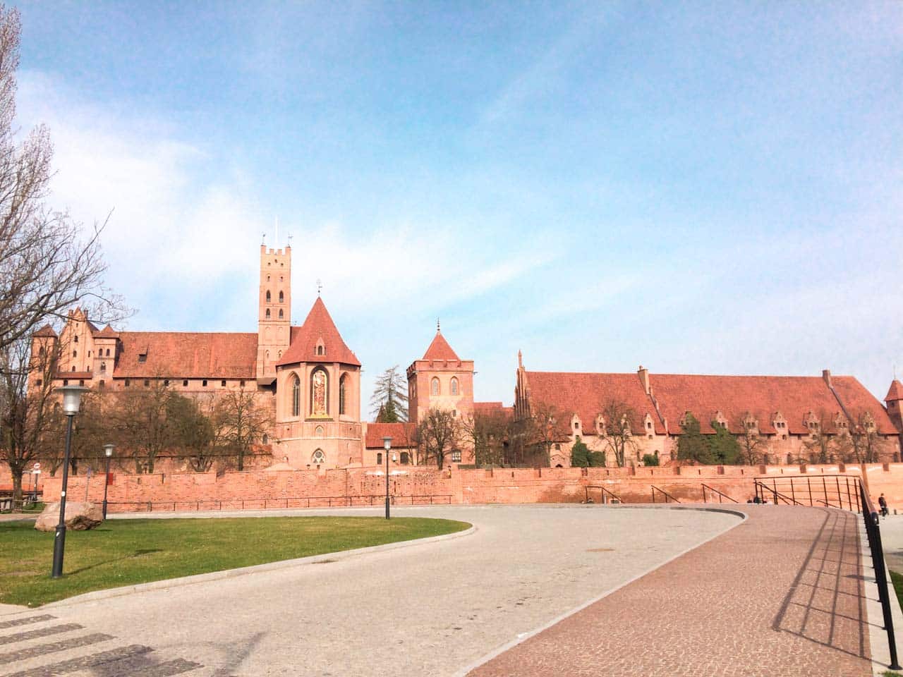 Wide view of Malbork Castle with red brick towers, tall walls, and a clear blue sky overhead
