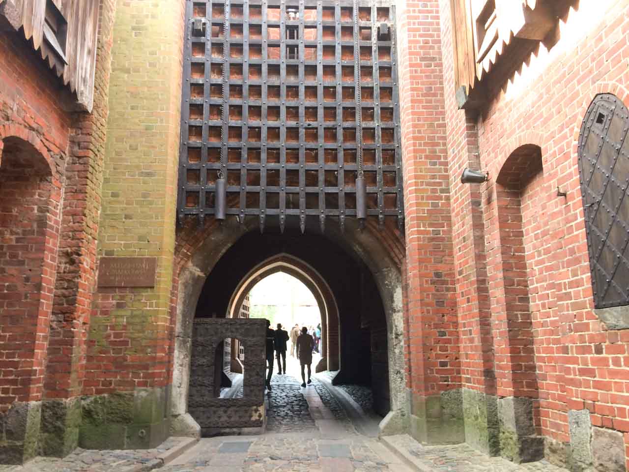 Brick gate tunnel with a heavy iron portcullis above, leading into Malbork Castle with people walking through
