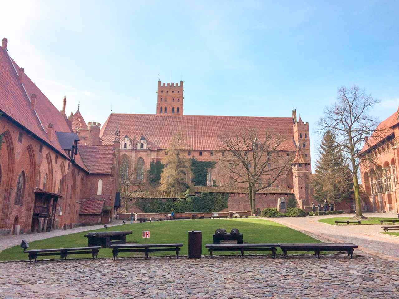 Malbork Castle courtyard with red brick buildings, a tall tower, and green lawns with old cannons