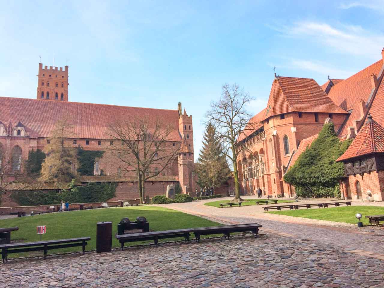 Main courtyard at Malbork Castle with benches, cannons, cobbled paths and red brick buildings covered in ivy
