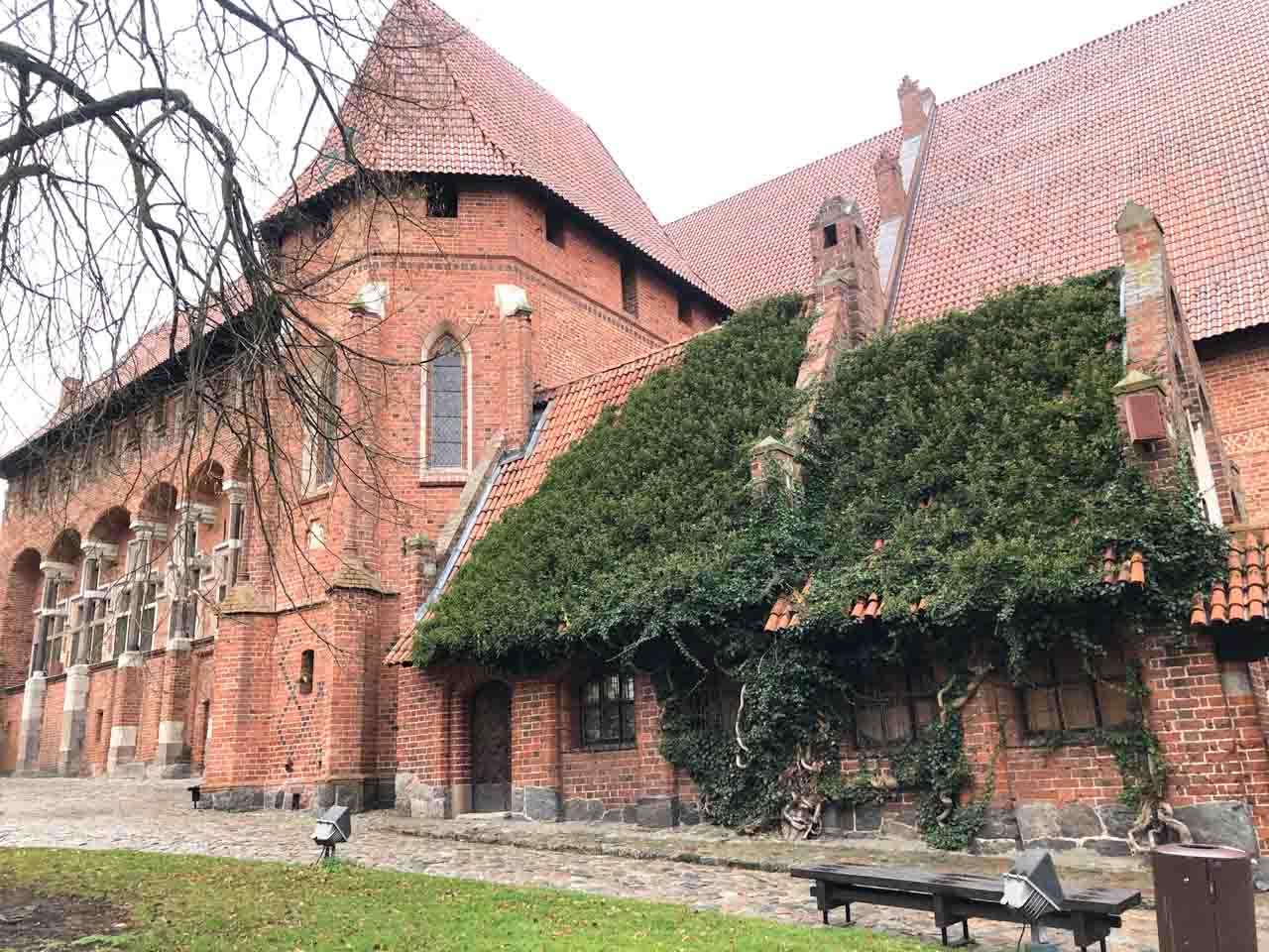 Red brick wing of Malbork Castle with tall chimneys and ivy covering the lower roof and windows, next to a cobbled path
