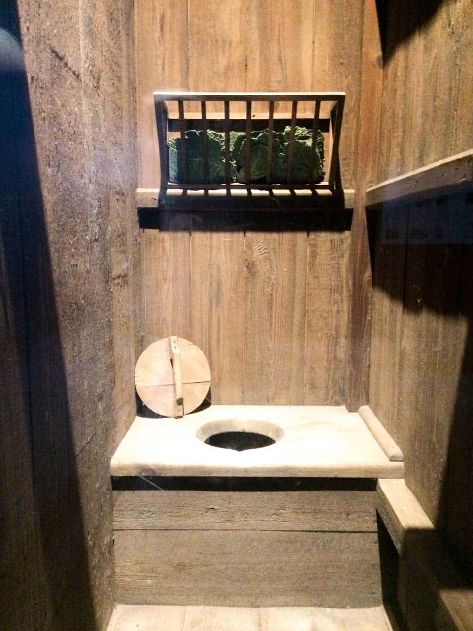 A medieval wooden toilet with a round hole, set in a small wooden cubicle with cabbage leaves stored on a shelf