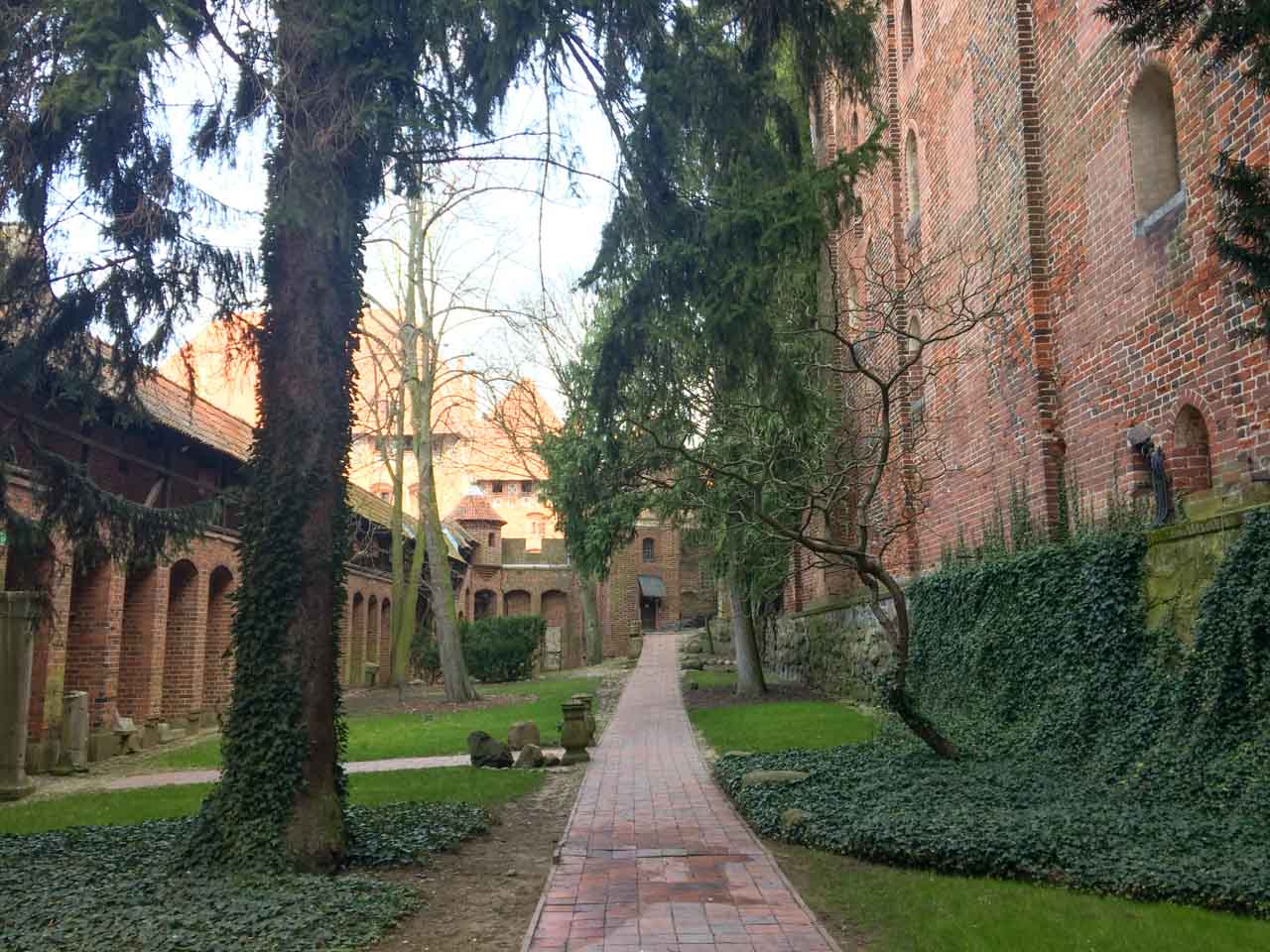 Peaceful garden path lined with ivy and trees, running alongside the red brick walls of Malbork Castle