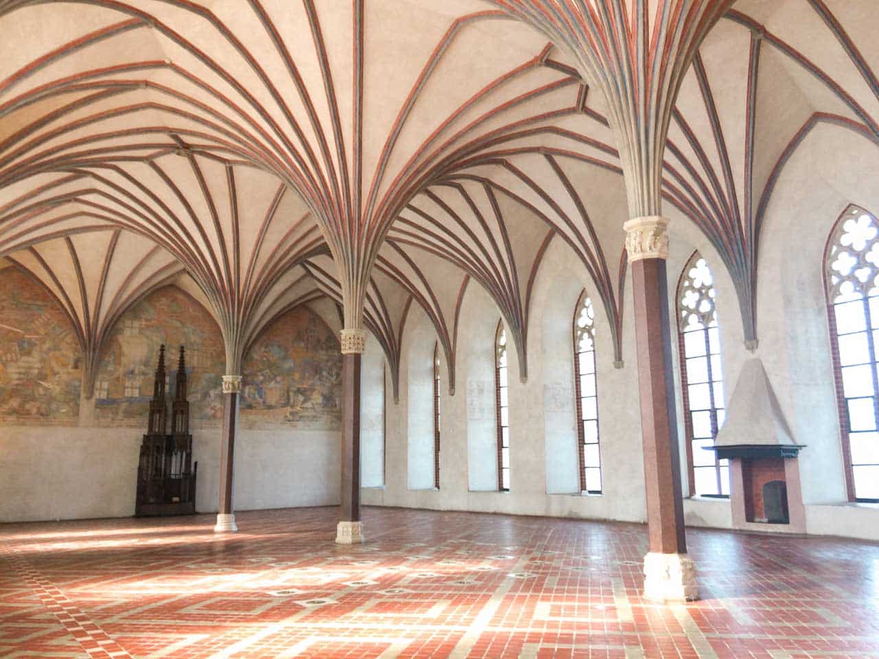 Light-filled hall inside Malbork Castle with a vaulted ceiling, tall windows, a wall mural, and a decorative fireplace