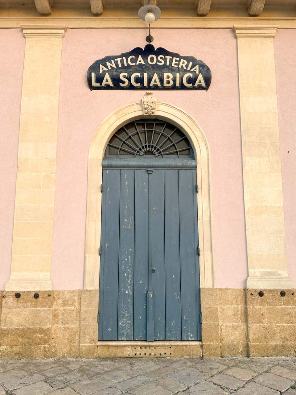 Blue wooden door of Antica Osteria La Sciabica in Brindisi, framed by a pink and cream wall with a sign above the archway