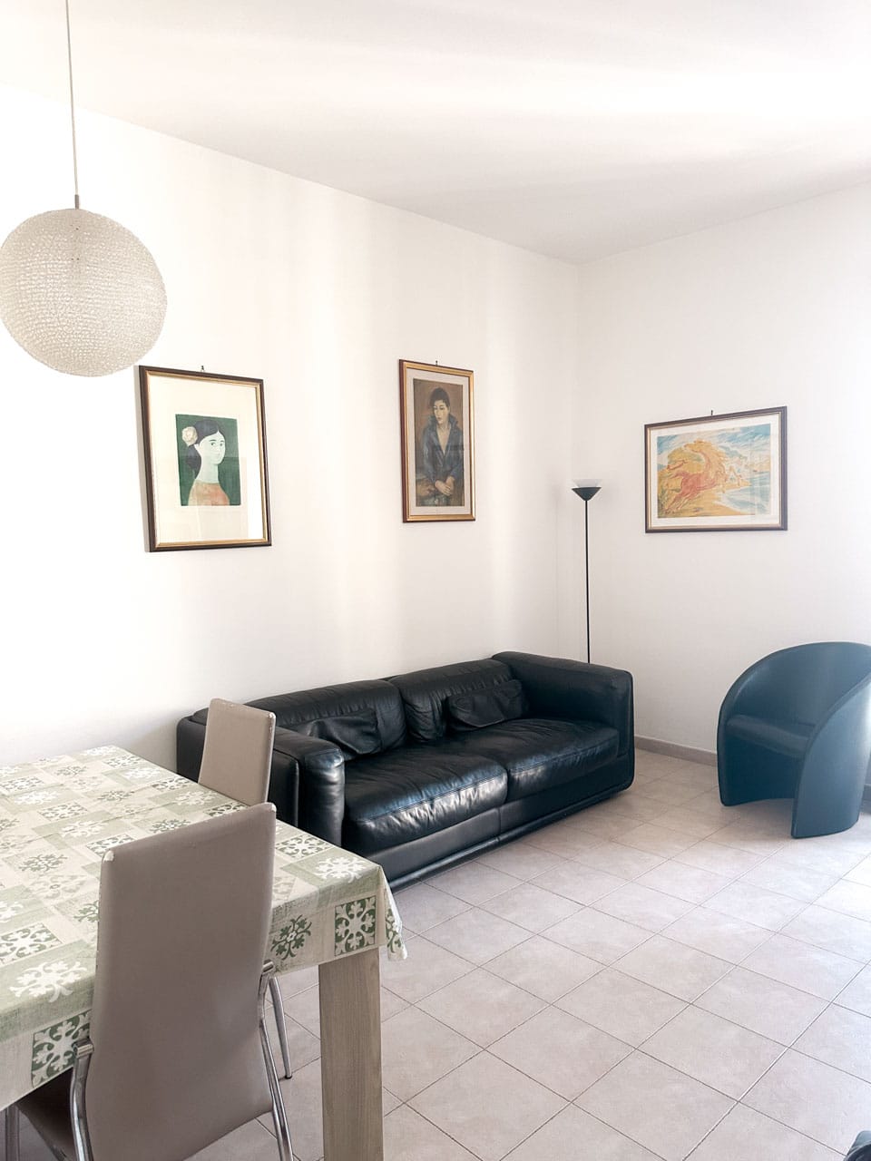 Bright living room with a black leather sofa, a black armchair, and framed art on white walls above tiled floors