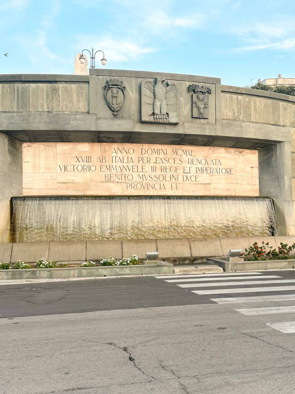 Fascist-era fountain in Brindisi with cascading water and a stone inscription featuring Mussolini's name
