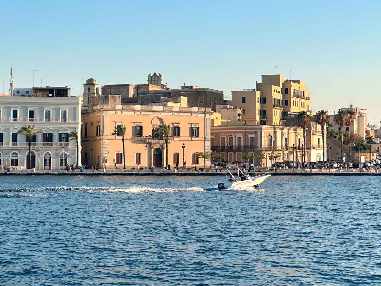 Speedboat gliding across the water with yellow and pink buildings along Brindisi's waterfront behind