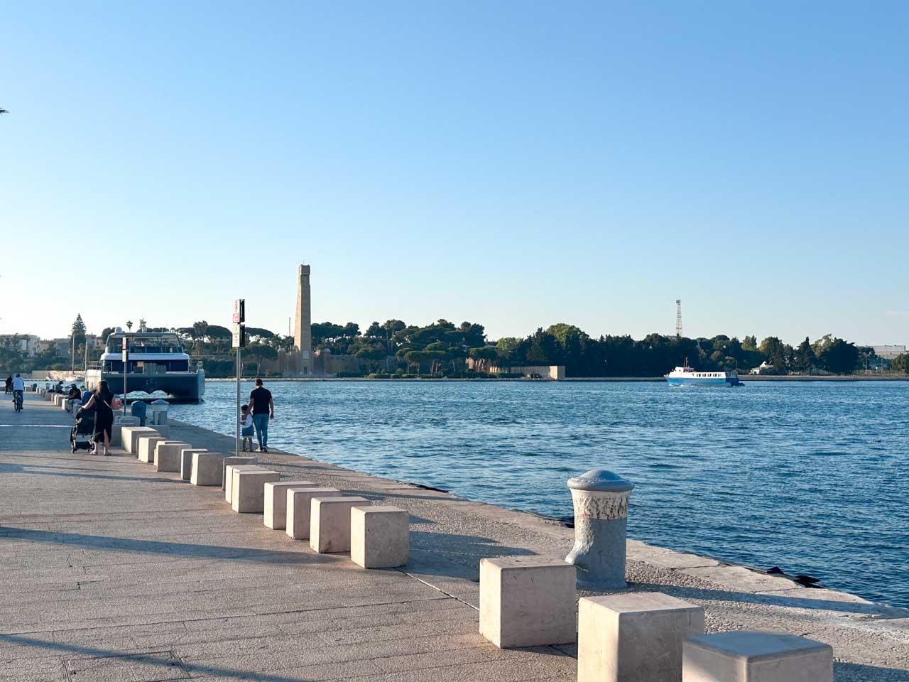 People walking and cycling along Brindisi’s waterfront promenade, with boats and the Monument to the Italian Sailor ahead